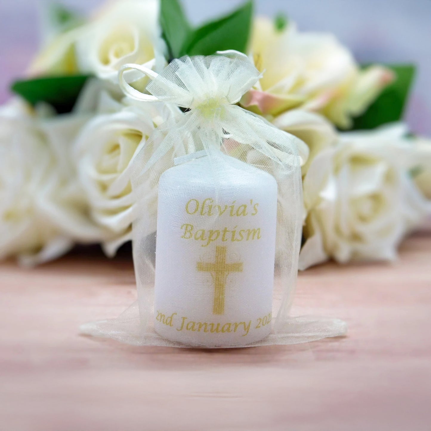A personalised white baptism candle tied with a sheer white organza bow, featuring gold text reading 'Olivia's Baptism' and a gold cross, set against a blurred background of white roses.