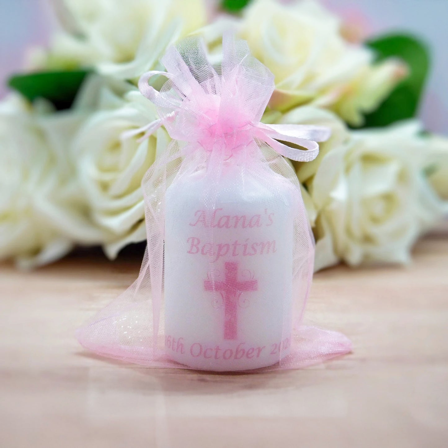 A small white  candle favour wrapped in sheer pink tulle with a tied bow, printed with the text 'Alana's Baptism' and a pink cross, set against a blurred background of white roses.
