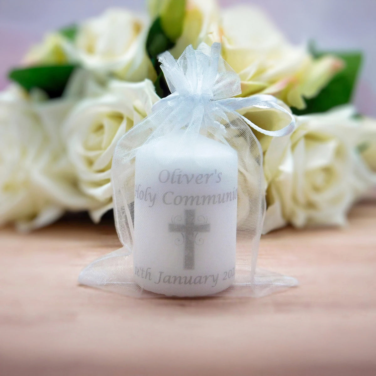A white candle wrapped in a sheer white organza bag with a ribbon, printed with the text 'Oliver's Holy Communion' and a grey cross, set against a blurred background of white roses.