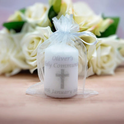 A white candle wrapped in a sheer white organza bag with a ribbon, printed with the text 'Oliver's Holy Communion' and a grey cross, set against a blurred background of white roses.