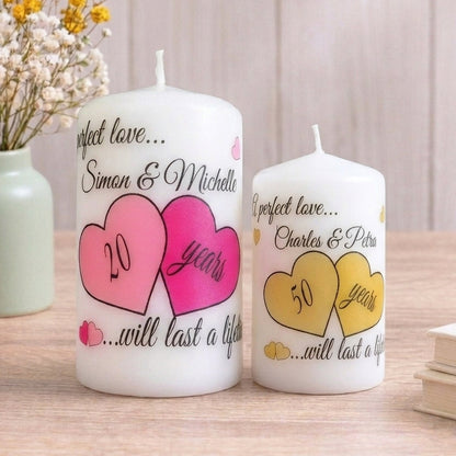 Two white pillar candles with custom printed heart designs and text, displayed on a wooden surface next to a vase of dried flowers and a stack of books.