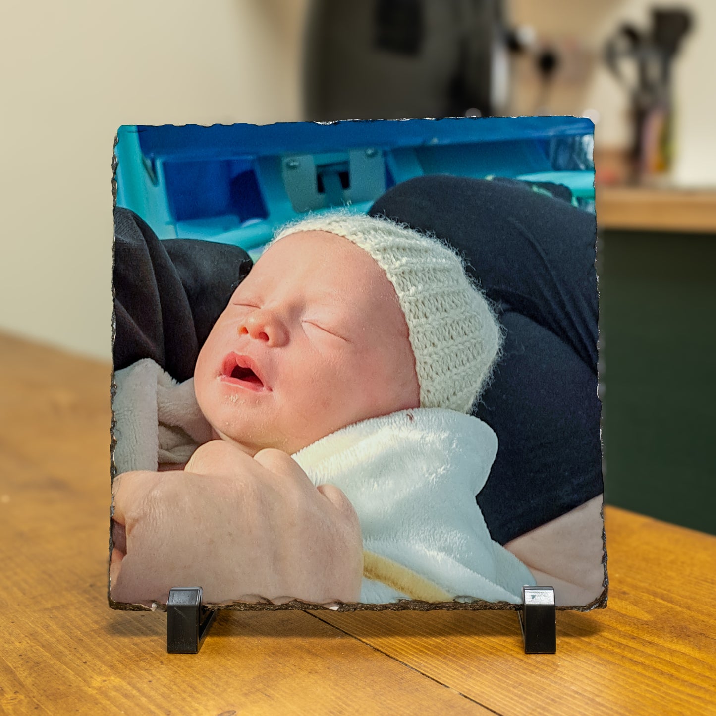A square slate photo plaque on a black metal stand, featuring a printed photograph of a sleeping newborn baby wearing a white knit hat, resting on a wooden table.