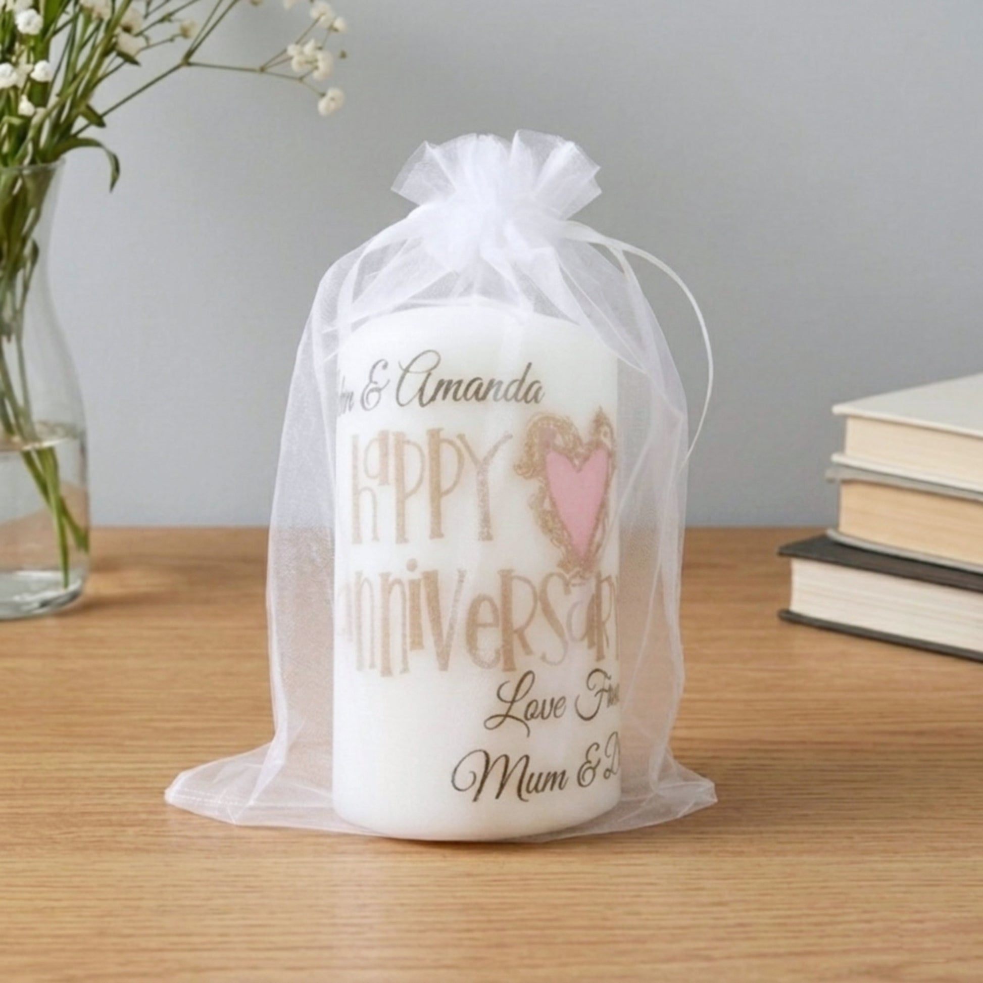 A white personalised candle wrapped in a sheer white organza bag, sitting on a wooden table. The candle features gold and pink text reading 'Happy Anniversary Love From Mum & Dad' with a gold heart outline.