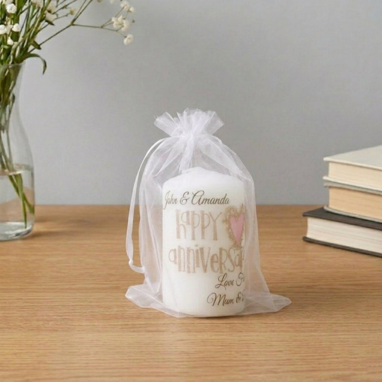 A personalised white anniversary candle inside a sheer white organza gift bag, displayed on a wooden table with a vase of flowers and books in the background.