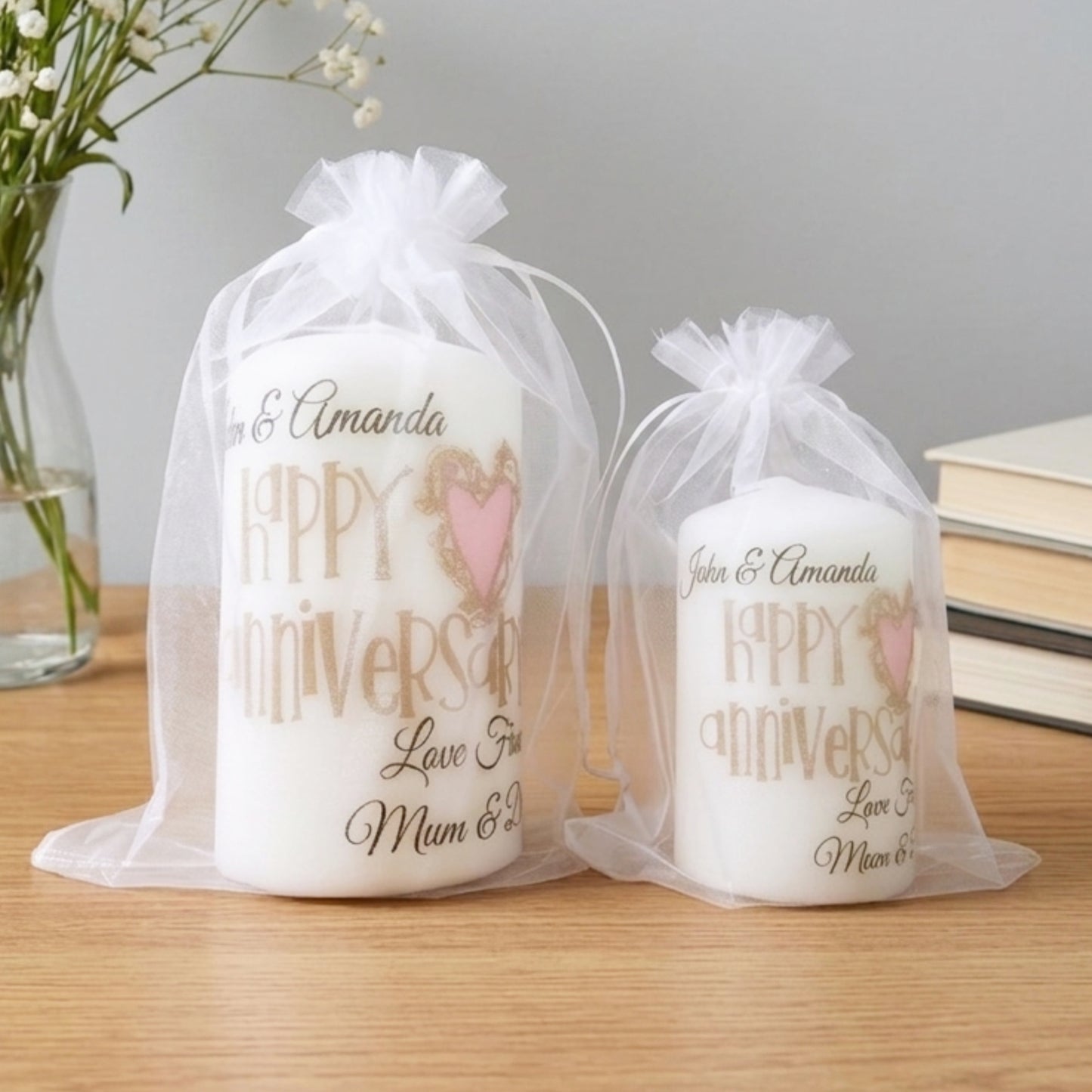 Two personalised white pillar candles in sheer white organza gift bags, displayed on a wooden tabletop with a vase of flowers and books in the background. The candles feature gold and pink text reading 'Happy Anniversary Love You Mum & Dad' with customizable names.