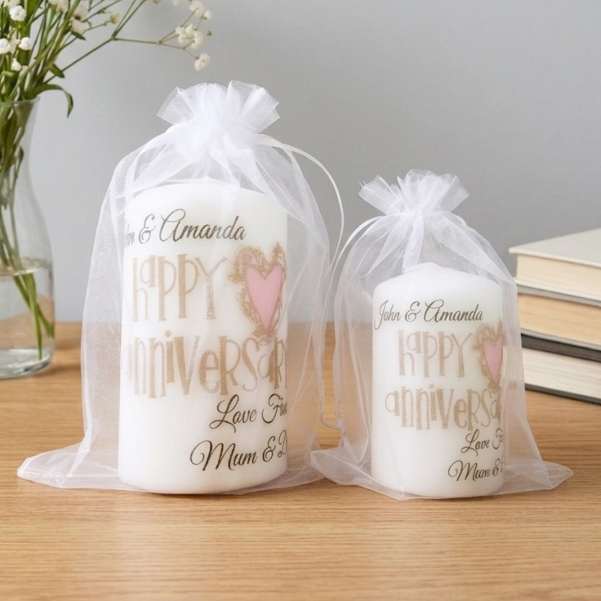 Two personalised white pillar candles in sheer white organza gift bags, displayed on a wooden tabletop with a vase of flowers and books in the background. The candles feature gold and pink text reading 'Happy Anniversary Love You Mum & Dad' with customizable names.