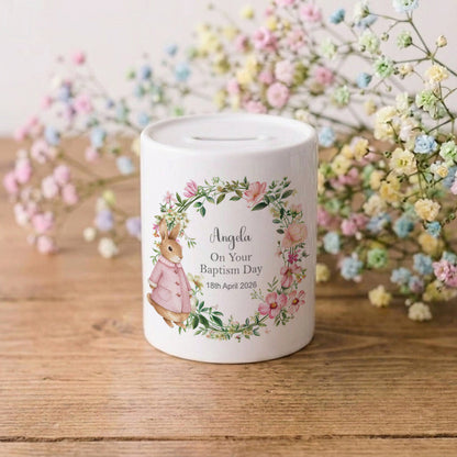 A white ceramic baby baptism money box with a personalised illustration of a bunny in a floral wreath, sitting on a wooden surface with baby's breath flowers in the background.
