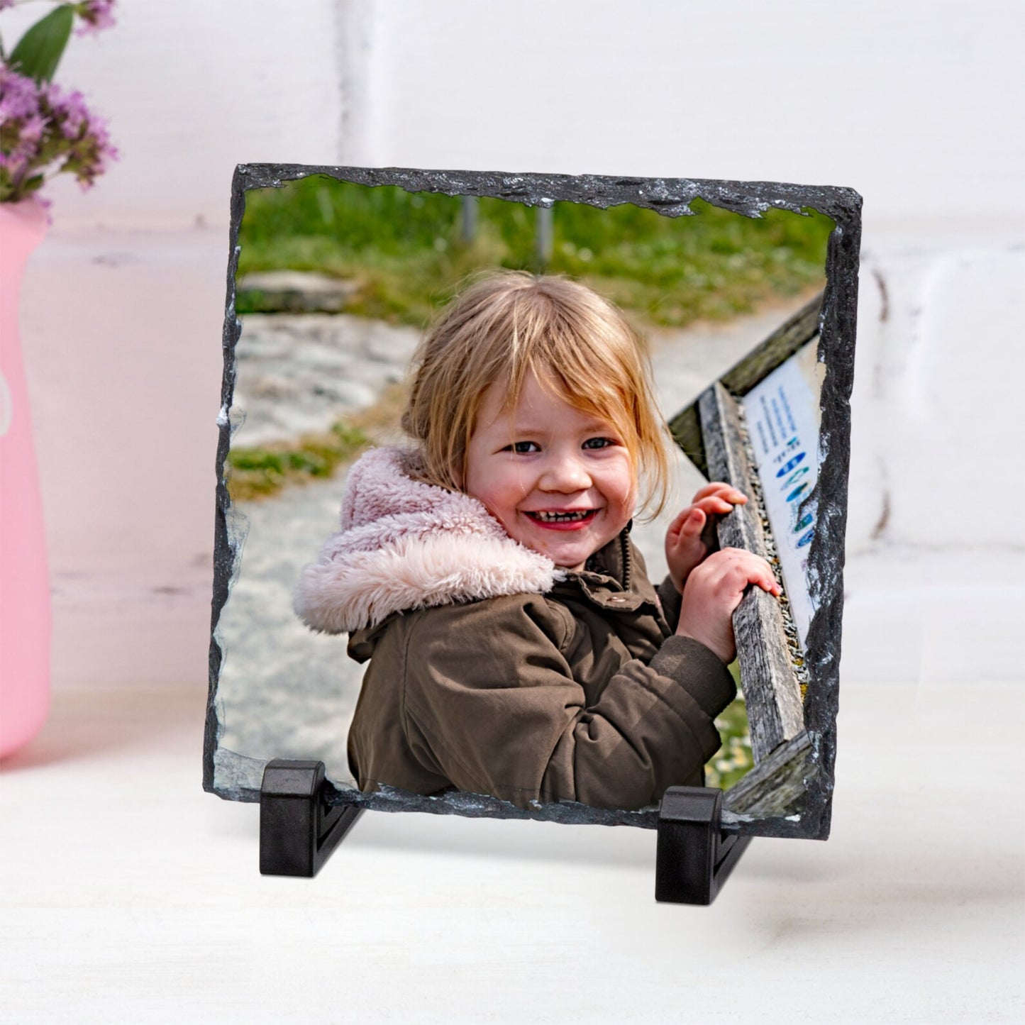 A square slate photo frame stand holding a custom photo of a smiling young girl in a brown jacket with a pink hood. The frame is displayed on a white surface against a white wall, with a pink vase of flowers partially visible on the left.