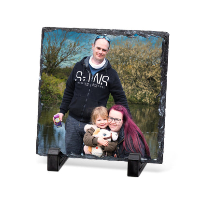A square slate photo plaque on a black stand, displaying a custom family portrait of a man, woman, and child standing by a lake. The plaque is shown on a clean white background.
