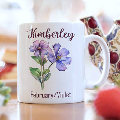 White mug with floral design and 'Kimberley' text, set against a blurred tea-making scene.