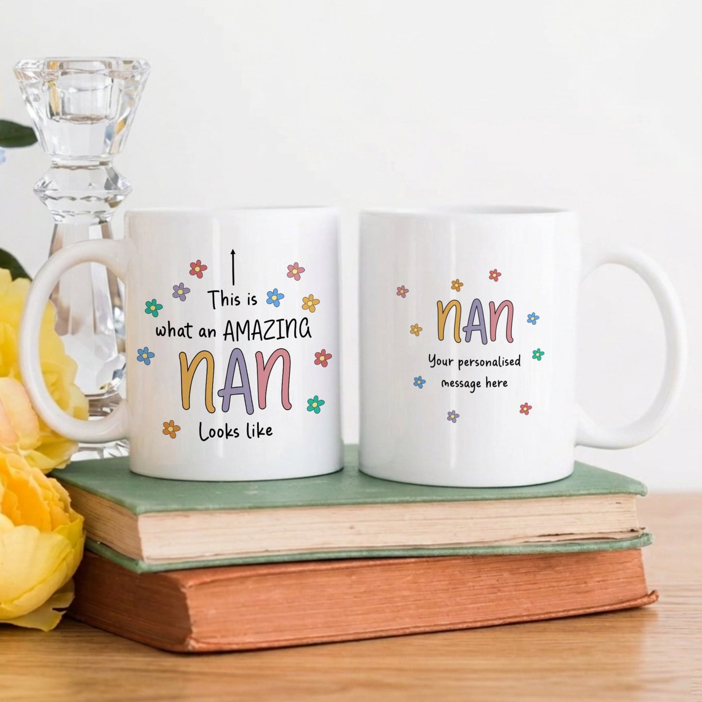 Two white ceramic mugs showing both sides of the 'This is what an Amazing Nan looks like' design with colourful floral details and a personalised message option, displayed on stacked books with yellow flowers