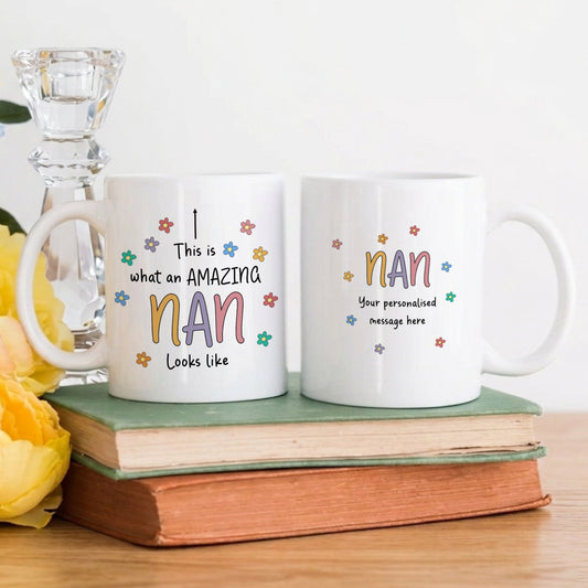 Two white ceramic mugs showing both sides of the 'This is what an Amazing Nan looks like' design with colourful floral details and a personalised message option, displayed on stacked books with yellow flowers