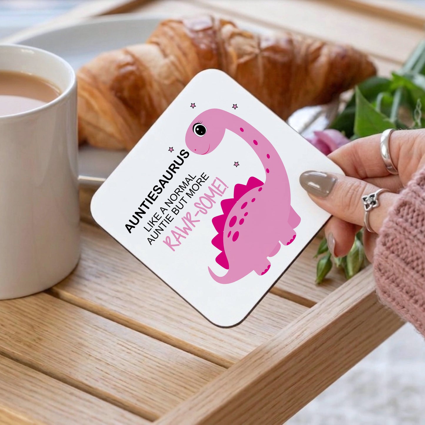 A square white coaster with rounded corners held by a hand, featuring a pink cartoon dinosaur and the text 'AUNTIES AURUS LIKE A NORMAL AUNTIE BUT MORE RAWR-SOME!'. The scene is set on a wooden tray with a cup of coffee, a croissant, and green leaves in the background.