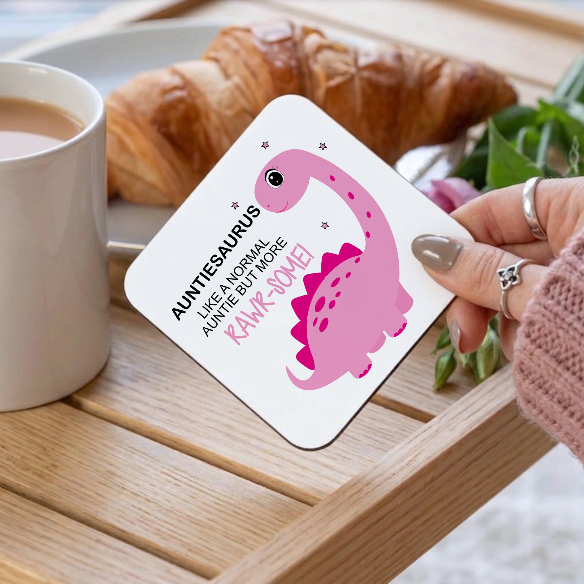 A square white coaster with rounded corners held by a hand, featuring a pink cartoon dinosaur and the text 'AUNTIES AURUS LIKE A NORMAL AUNTIE BUT MORE RAWR-SOME!'. The scene is set on a wooden tray with a cup of coffee, a croissant, and green leaves in the background.