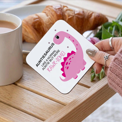 A square white coaster with rounded corners held by a hand, featuring a pink cartoon dinosaur and the text 'AUNTIES AURUS LIKE A NORMAL AUNTIE BUT MORE RAWR-SOME!'. The scene is set on a wooden tray with a cup of coffee, a croissant, and green leaves in the background.
