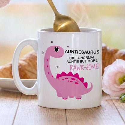 A white ceramic mug with a pink dinosaur graphic and the text 'AUNTIESAURUS LIKE A NORMAL AUNTIE BUT MORE RAWK-SOME!' sits on a wooden table. A hand holding a gold spoon stirs the mug, with a blurred breakfast scene in the background.