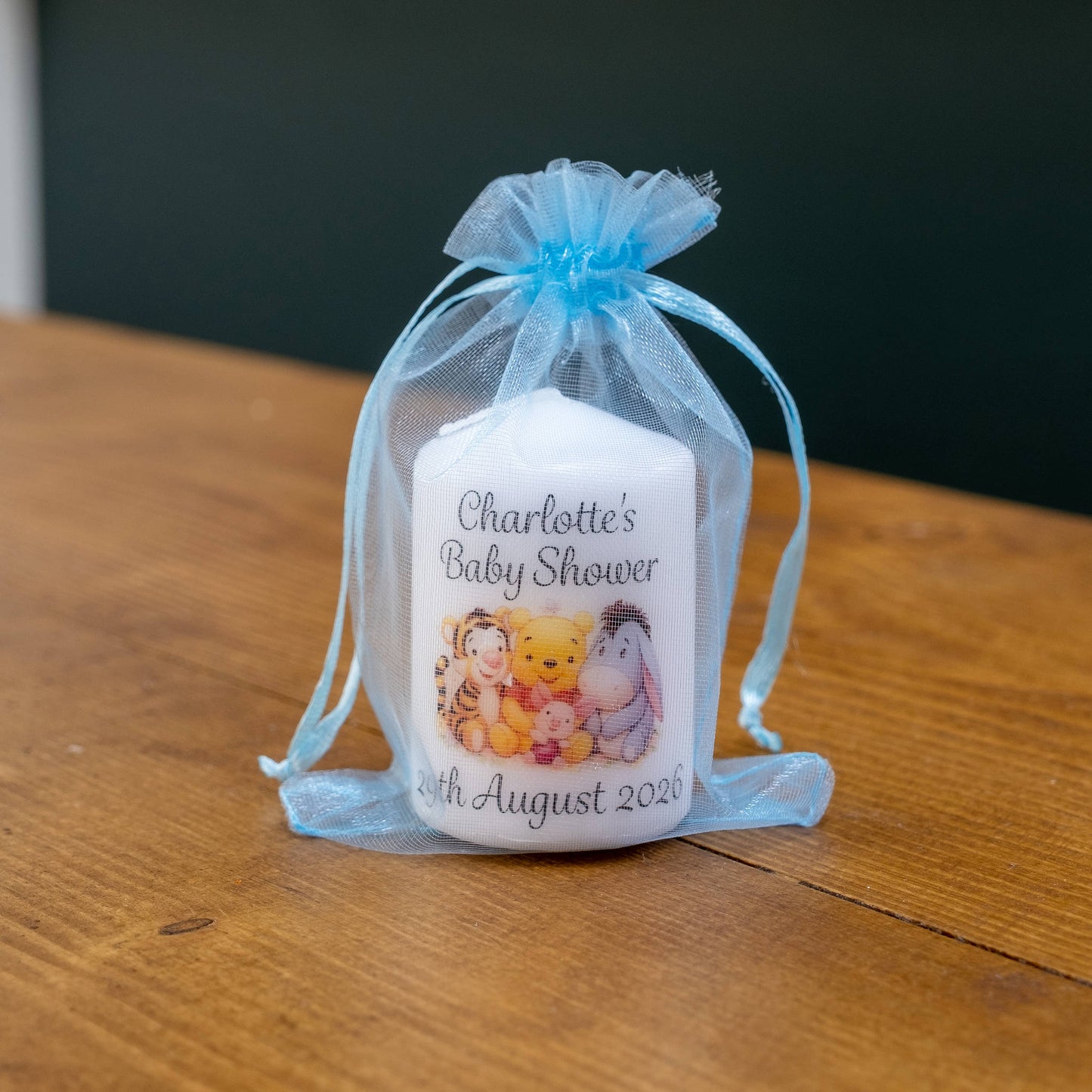 A personalised white candle with a light blue sheer organza drawstring pouch, containing a white candle printed with 'Charlotte's Baby Shower' and Winnie the Pooh characters, set on a wooden table.