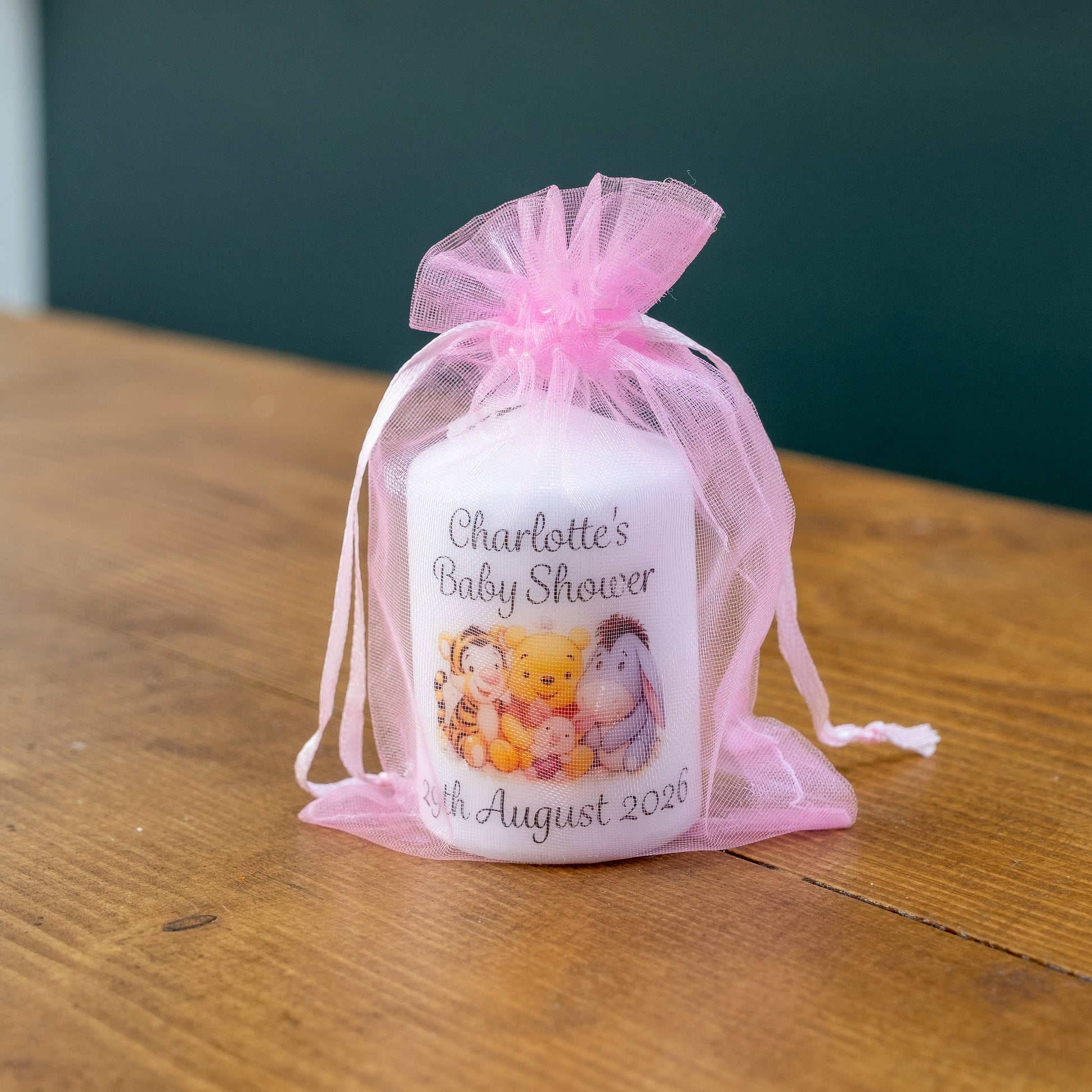 A personalised baby shower candle in a white glass jar, wrapped in a pink tulle organza bag and placed on a wooden table. The jar is printed with the text 'Charlotte's Baby Shower' and the date '15th August 2026', along with a cartoon character illustration featuring Winnie the Pooh and Tigger.