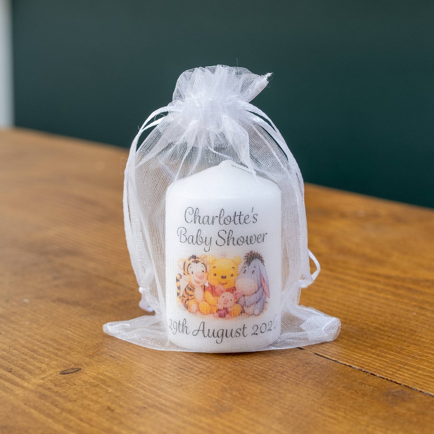 A personalised white baby shower candle wrapped in a sheer white organza bag, sitting on a wooden tabletop. The candle features custom printed text reading 'Charlotte's Baby Shower 15th August 202' and a colorful illustration of Winnie the Pooh characters.