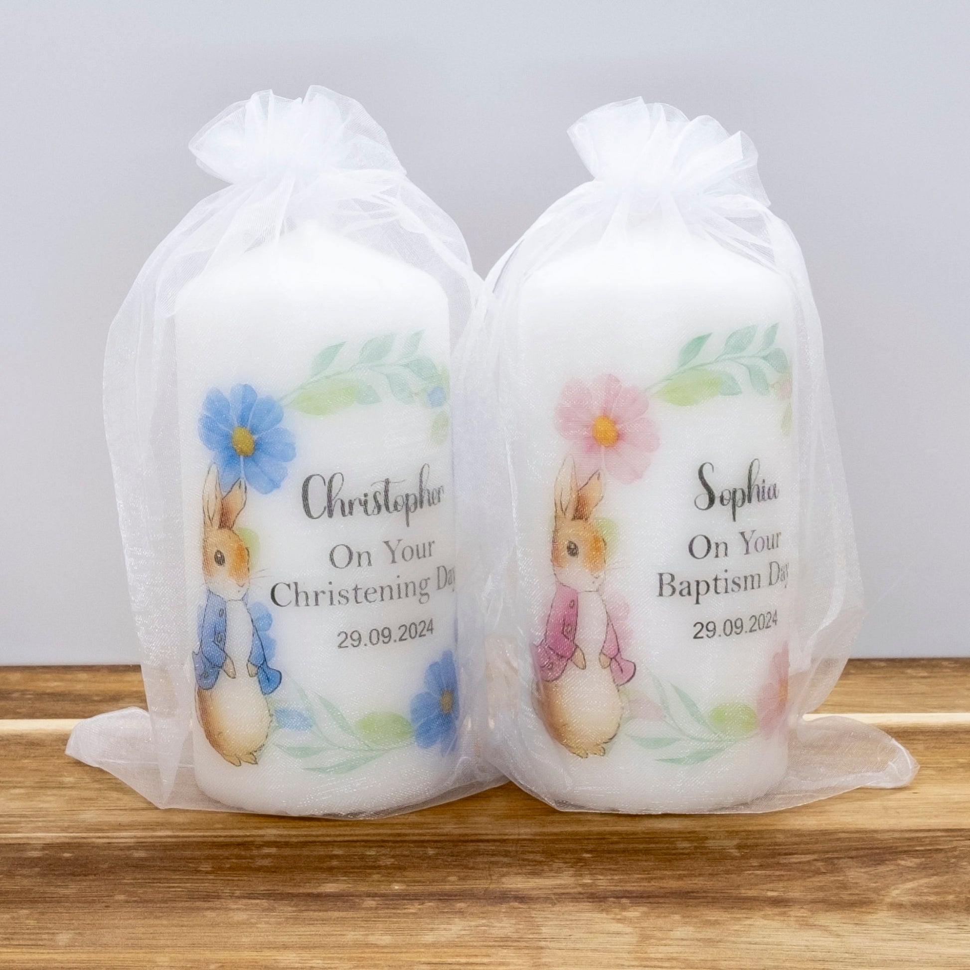 Two personalised baby baptism candles in clear organza bags, one labelled Christopher with blue flowers and one labelled Sophia with pink flowers, sitting on a wooden surface against a light grey background.