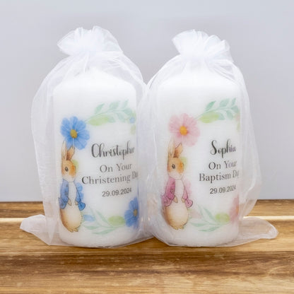Two personalised baby baptism candles in clear organza bags, one labelled Christopher with blue flowers and one labelled Sophia with pink flowers, sitting on a wooden surface against a light grey background.