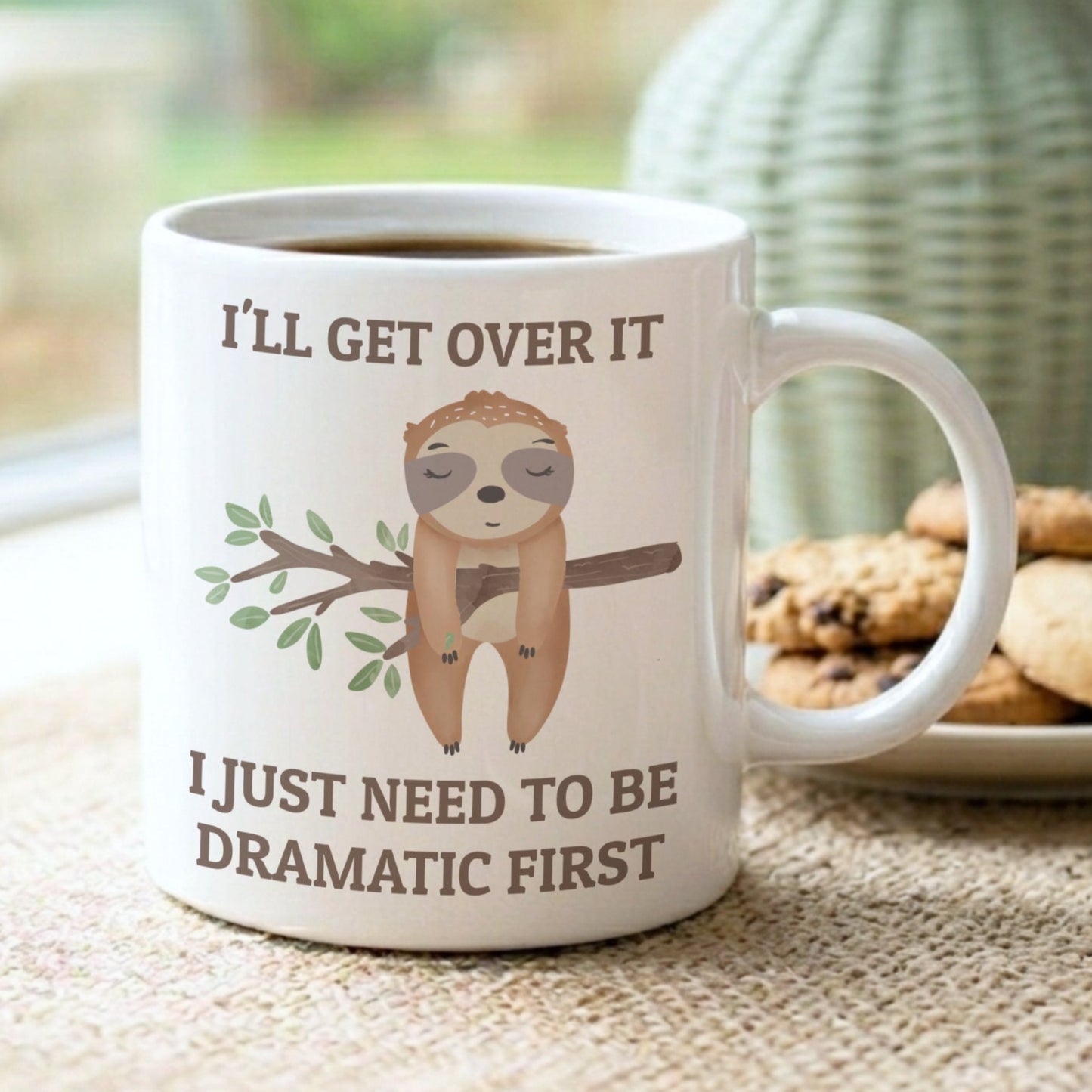 Mug with sloth illustration and humorous text on a table with cookies and a vase.