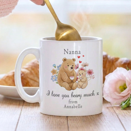 White mug with teddy bear design and text, held by a hand with a gold spoon.