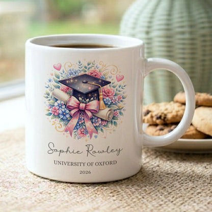 Personalised graduation mug with a floral mortarboard and pink bow design, customised with 'Sophie Rowley, University of Oxford, 2026', displayed on a windowsill with cookies and a sage green vase