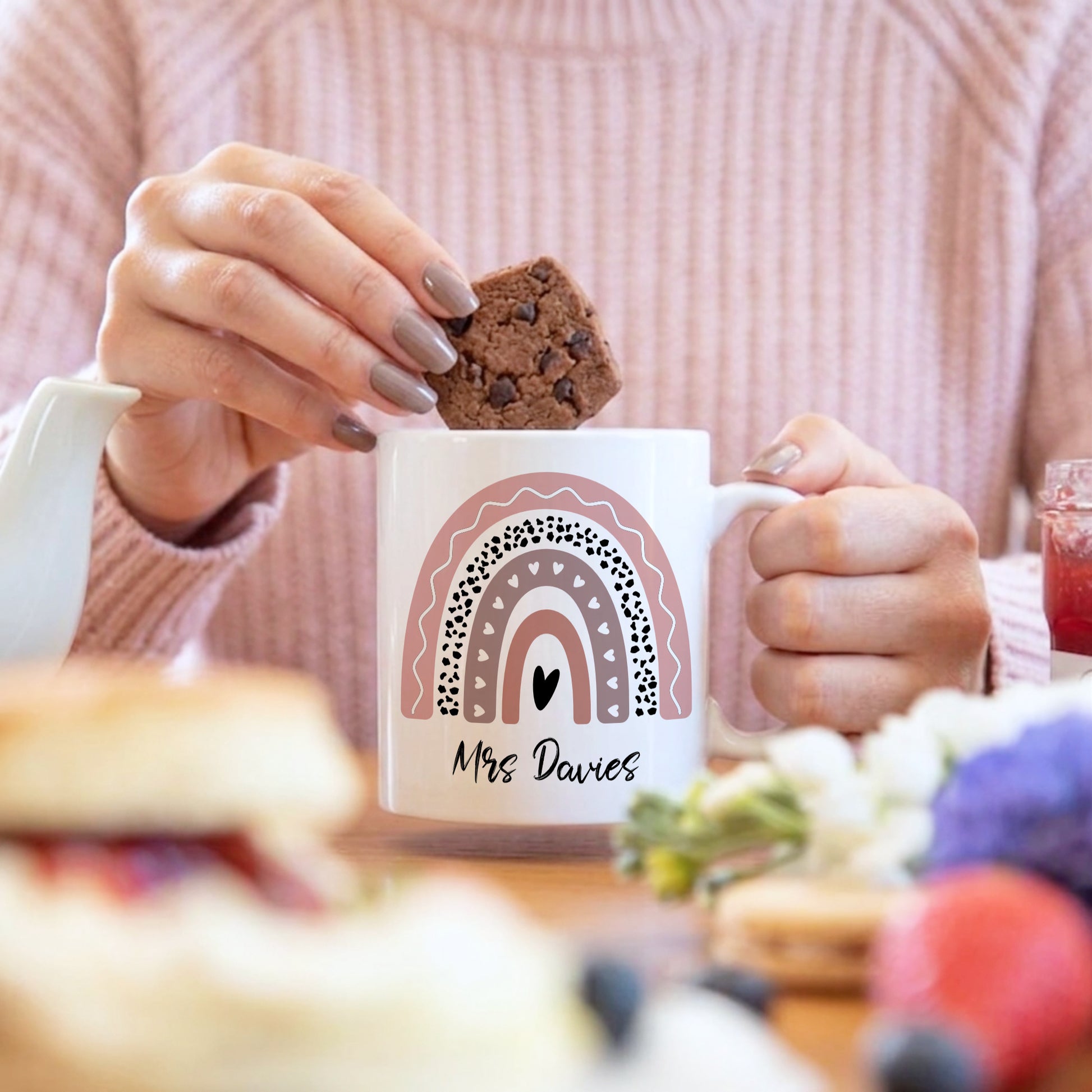 Person holding a mug with a rainbow design and 'Mrs Davies' text, eating a cookie.