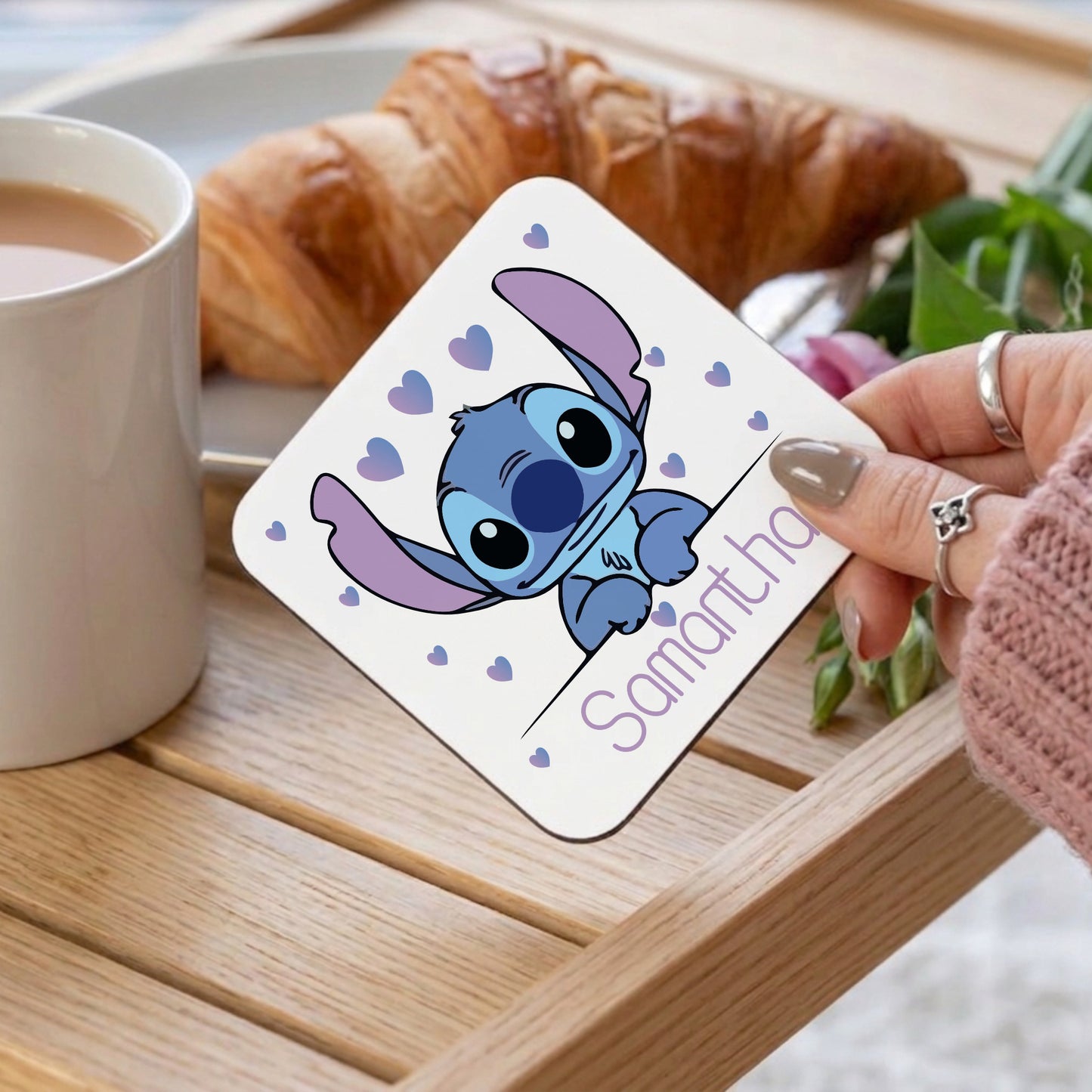 Personalised Stitch coaster with blue heart details and 'Samantha' name, being held over a wooden tray alongside a cup of tea and a croissant