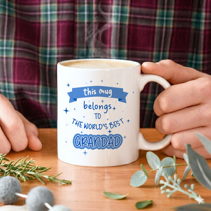 A man holding a white mug with "this mug belongs to the World's best Grandad" text in blue 
