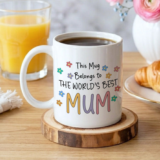 White mug with 'World's Best Mum' text on a wooden coaster, surrounded by coffee, juice, and pastries.