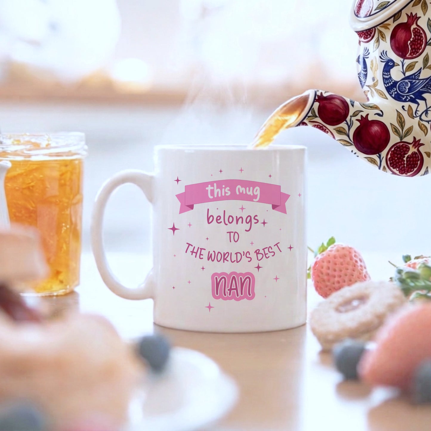 A white ceramic mug with pink text reading 'this mug belongs to THE WORLD'S BEST Nan' sits on a table with a floral teapot pouring tea into it, surrounded by breakfast items like a strawberry, donuts, and honey in a bright kitchen setting.