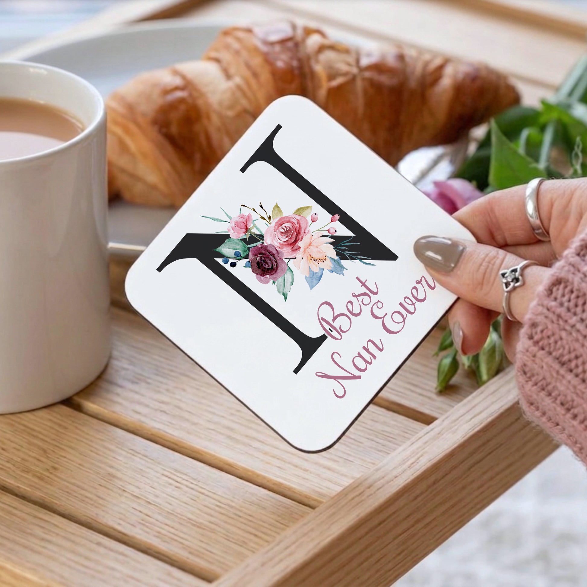 A hand holds a white square coaster featuring a large black letter N, pink script text reading 'Best Nan Ever,' and a floral illustration, set on a wooden tray with a mug, croissant, and flowers in the background.