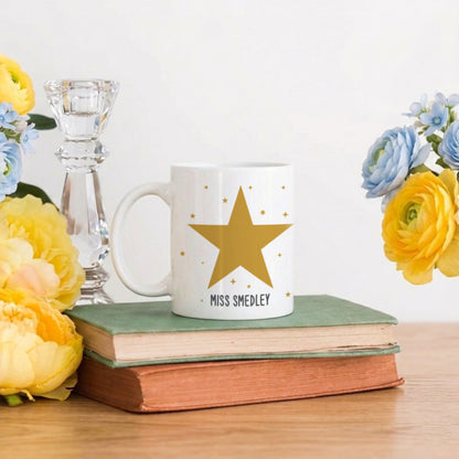 White mug with a gold star and text, placed on books with flowers and a candle in the background