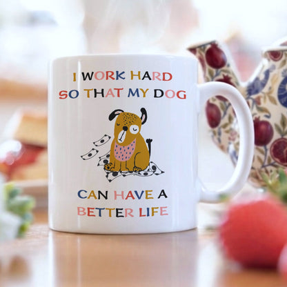 White ceramic mug with 'I Work Hard So That My Dog Can Have a Better Life' quote and cute illustrated dog surrounded by money, displayed on a table with a floral teapot and fresh strawberries