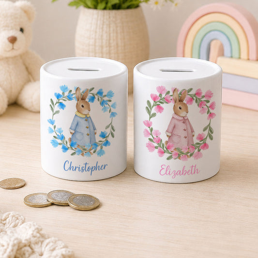 Two personalised white ceramic pig savings banks with Peter Rabbit illustrations, one in blue and one in pink, displayed on a wooden surface with coins and toys in the background.