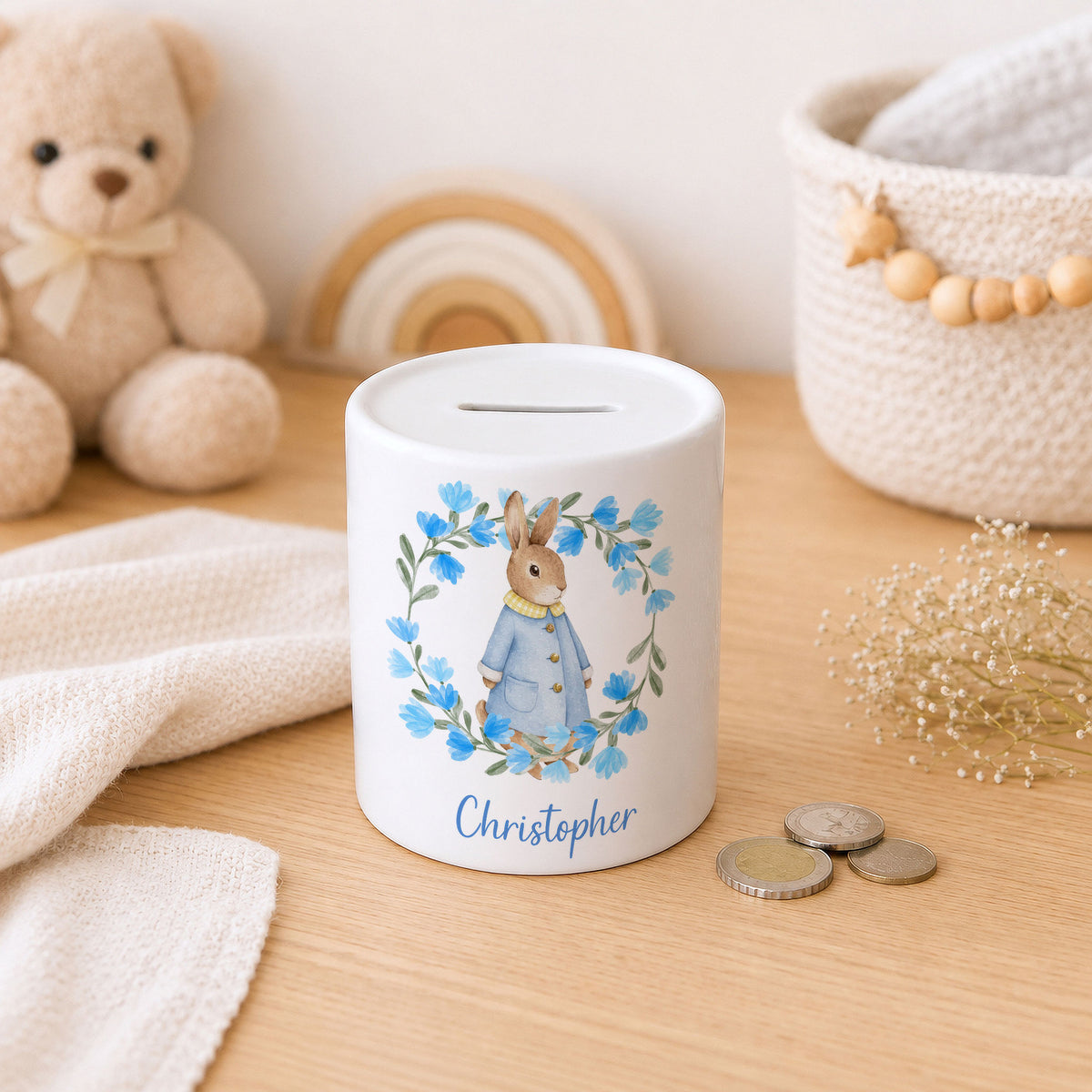 A white ceramic pig savings bank with a personalised name 'Christopher' and an illustration of a rabbit in a blue outfit surrounded by blue flowers. The bank is styled on a wooden table with a teddy bear, a woven basket, and coins in the background.