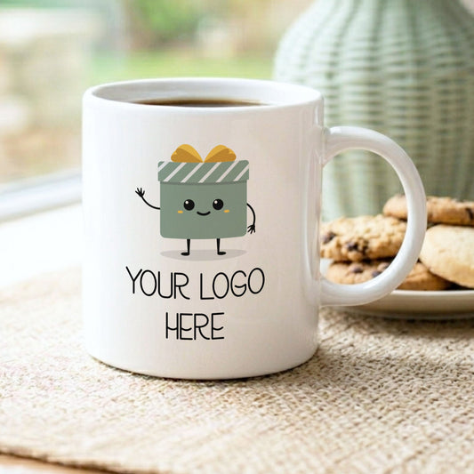 A white ceramic mug filled with coffee sits on a woven placemat by a window, with a plate of cookies and a green vase blurred in the background. The mug has a cartoon gift box character printed on it and the text 'YOUR LOGO HERE'.