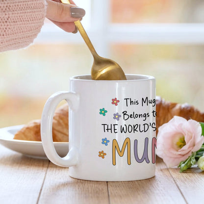 White mug with colorful text and flowers, held by a hand with a gold spoon, on a wooden table with a blurred background.
