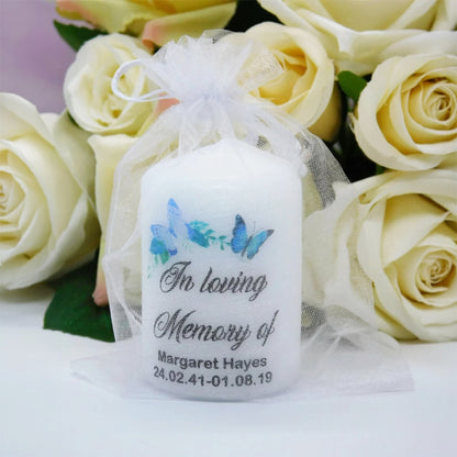 A personalised white taper candle wrapped in a sheer white organza bag, decorated with blue butterflies and the text 'In loving Memory of Margaret Hayes' with a date. The candle is styled in front of a bouquet of cream-coloured roses.