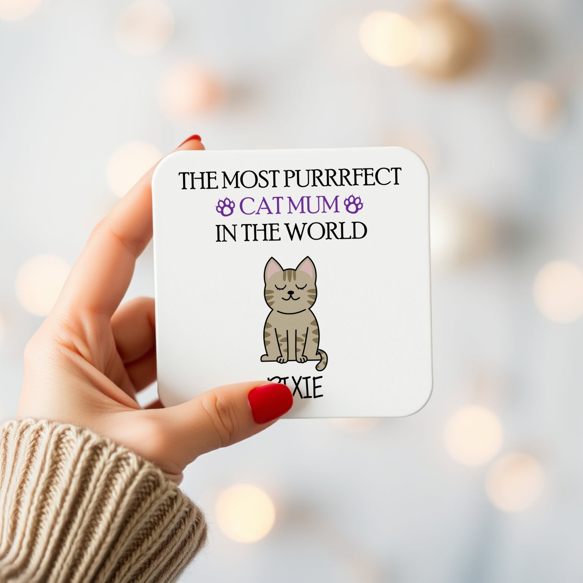 Coaster with a cat illustration and text held by a hand against a blurred light background