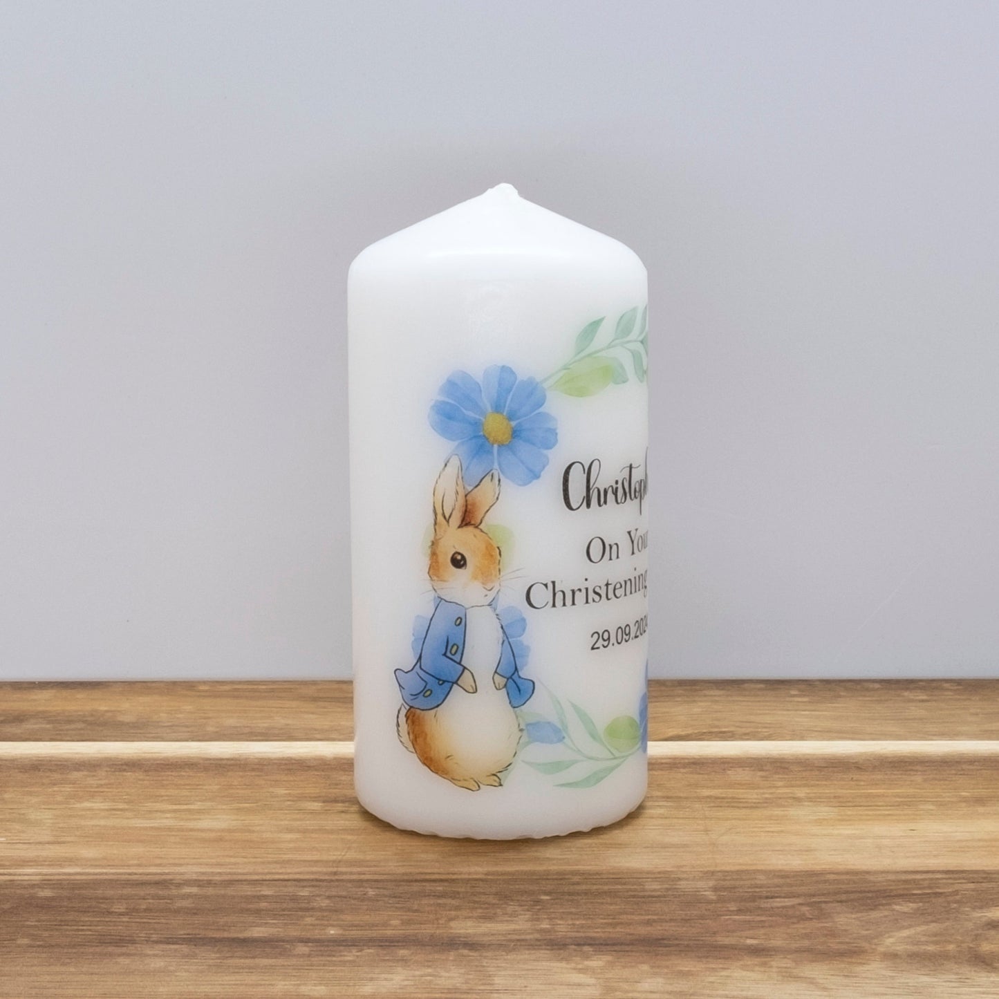 A white cylindrical candle with a pointed top, decorated with a Peter Rabbit illustration and blue floral details, standing on a wooden surface against a light grey background.