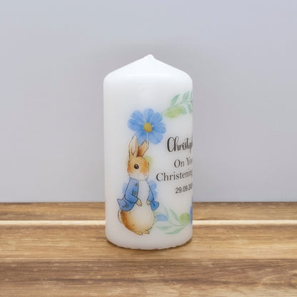 A white cylindrical candle with a pointed top, decorated with a Peter Rabbit illustration and blue floral details, standing on a wooden surface against a light grey background.