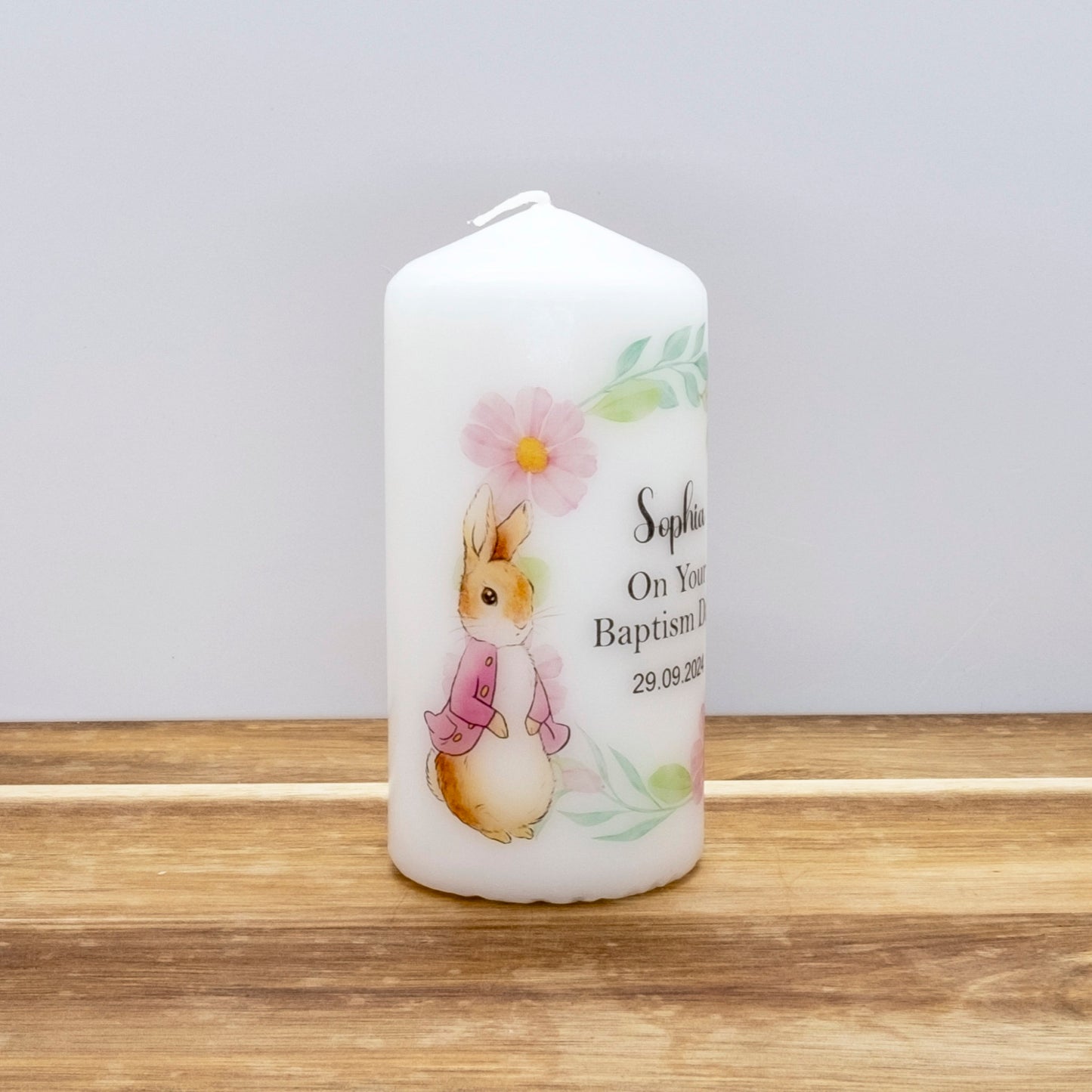 A white personalised birthday candle featuring a watercolour illustration of Beatrix Potter's Peter Rabbit and a pink flower, with custom printed text on a wooden surface against a light grey background.