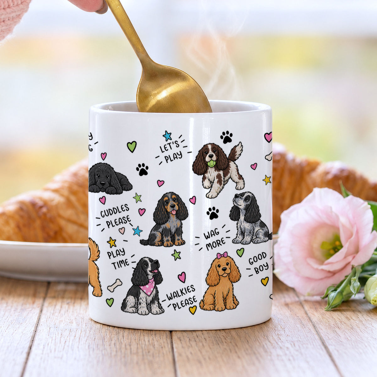A white ceramic coffee mug with a cute cartoon dog pattern, shown on a wooden table with a gold spoon in, a croissant in the background, and a pink flower in the foreground.