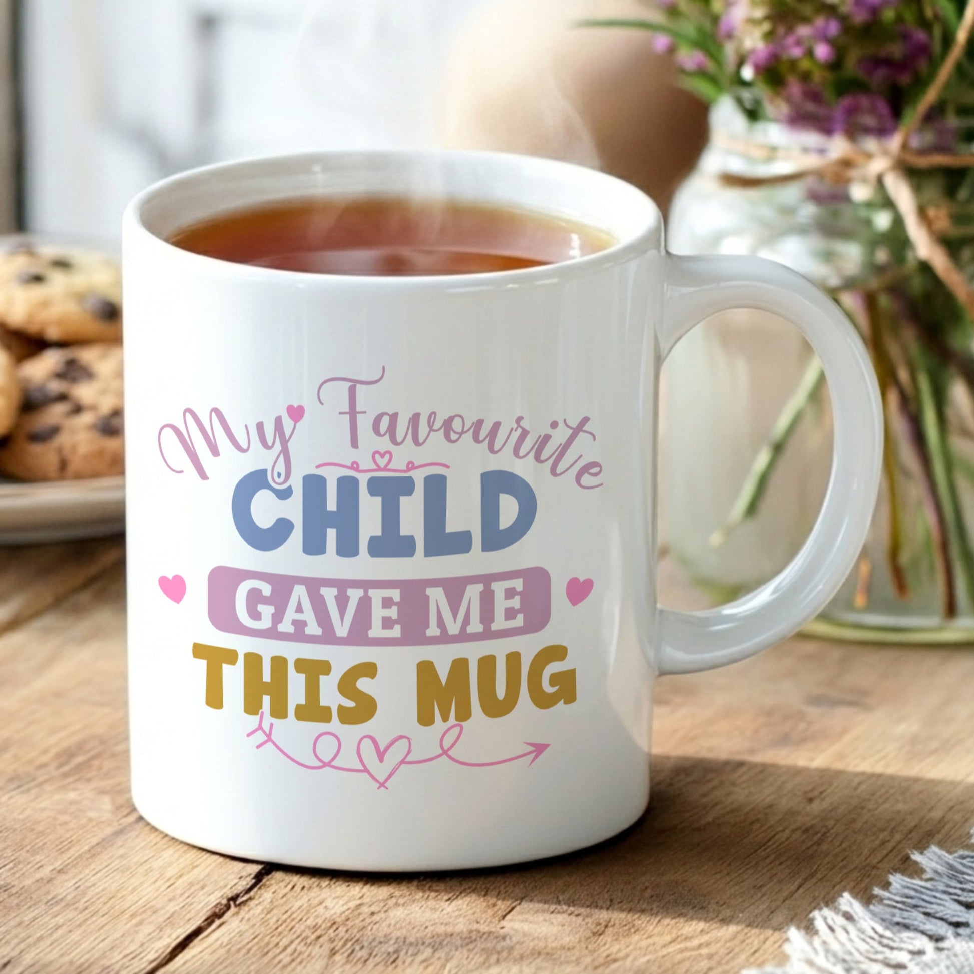 White 'My Favourite Child Gave Me This Mug' mug filled with tea, displayed on a wooden table with chocolate chip cookies and a vase of purple wildflowers