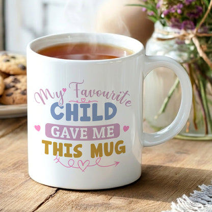 White 'My Favourite Child Gave Me This Mug' mug filled with tea, displayed on a wooden table with chocolate chip cookies and a vase of purple wildflowers