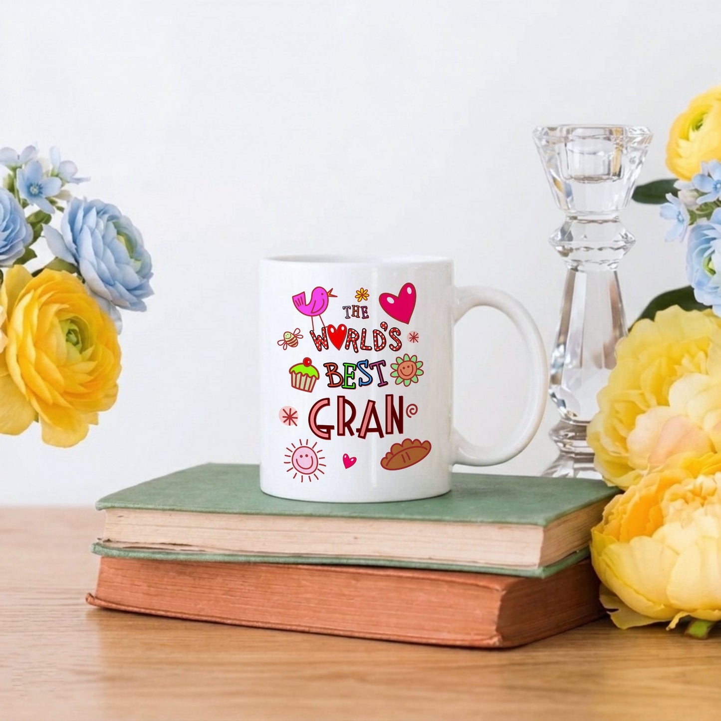 White mug with colorful text on a stack of books surrounded by flowers and a candle.
