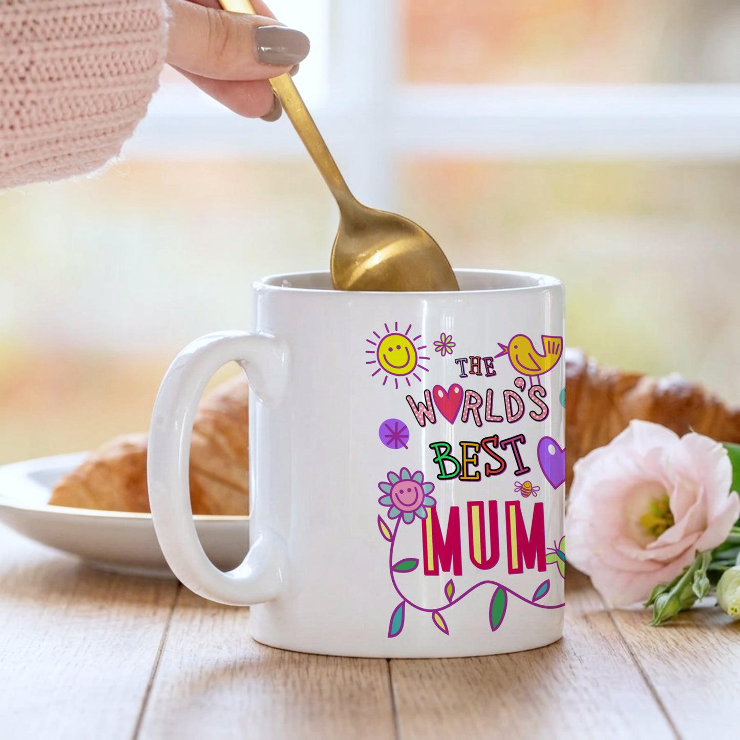 White mug with 'The World's Best Mum' text and colorful graphics, being stirred with a gold spoon.